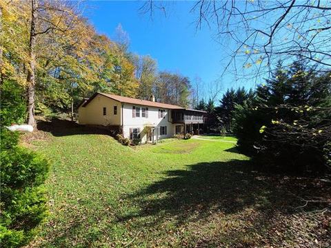 The two-story house seen across a sloping side lawn, with light siding, dark shutters and autumn trees behind it.