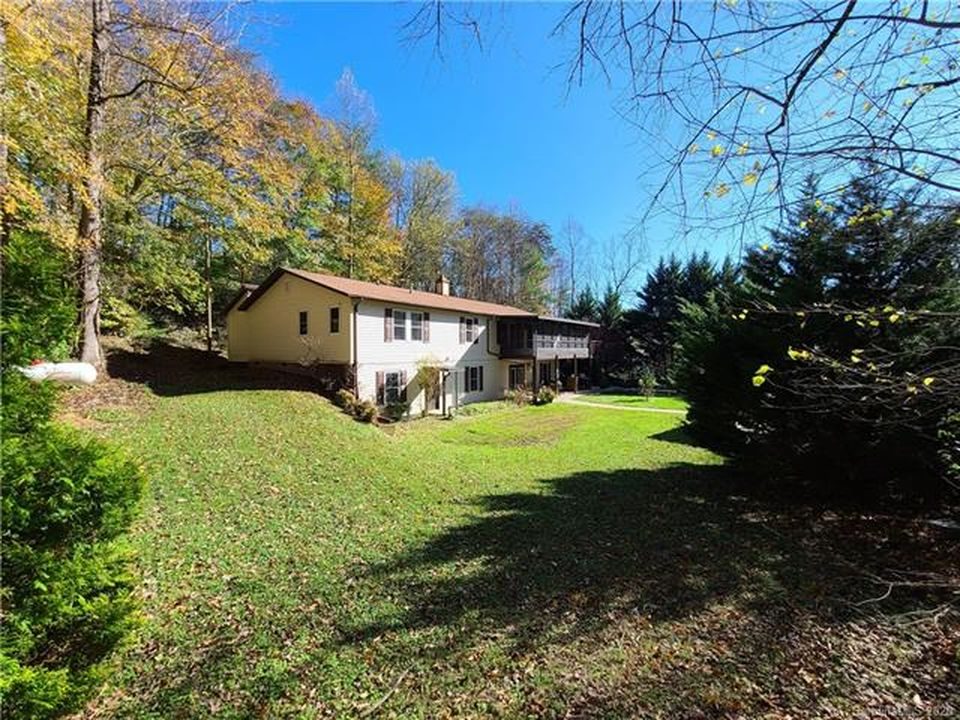 The two-story house seen across a sloping side lawn, with light siding, dark shutters and autumn trees behind it.