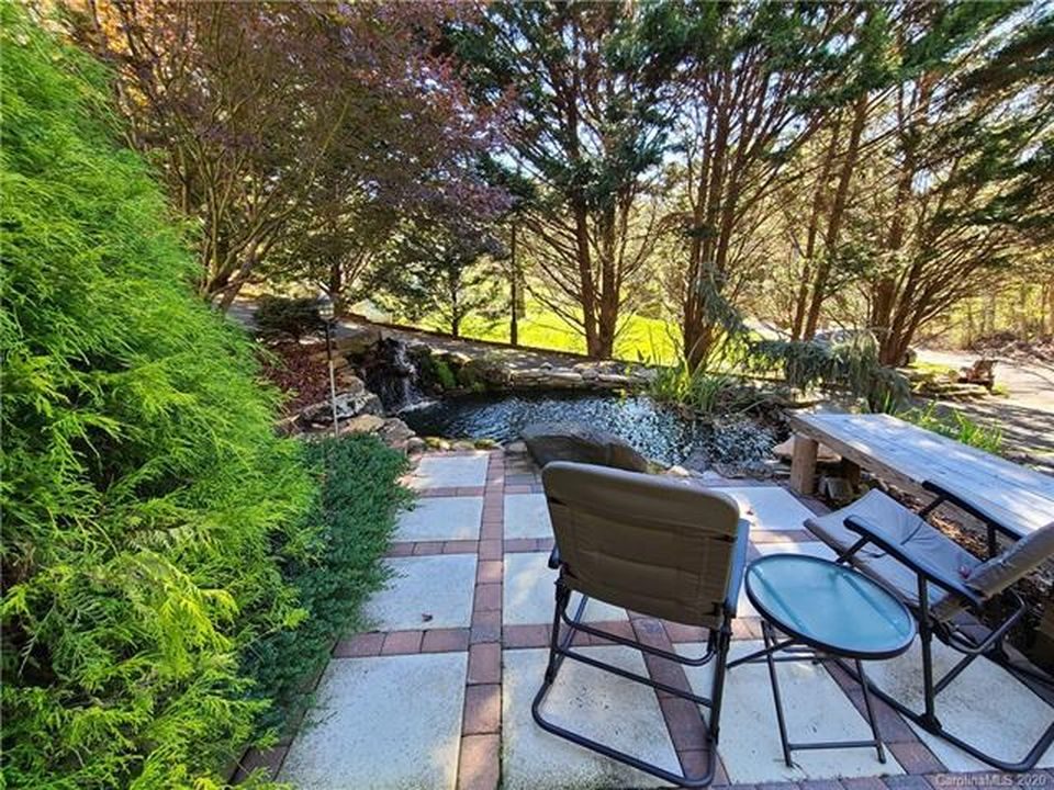 Paver patio with chairs and a small table beside a waterfall pond, framed by evergreens.